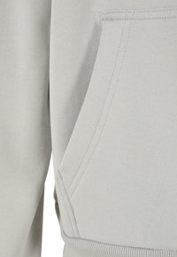 Gros plan sur un tissu gris clair montrant une poche kangourou avec des coutures visibles et un ourlet côtelé d'un sweat-shirt ou d'un sweat à capuche.