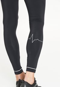 Schwarze Sportleggings aus dehnbarem Material, mit einem weißen Herzschlaglinien-Design an der Wade und weißen Akzentnähten am Saum.