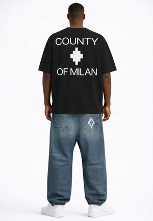 Mężczyzna noszący czarną, za dużą koszulkę z napisem „County of Milan” i symbolem, w połączeniu z luźnymi niebieskimi dżinsami z tym samym symbolem na tylnej kieszeni.