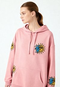 Rosa hoodie med en framficka, med broderi av sol och måne i blått och gult på ärmarna. Mjukt tyg, avslappnad design.