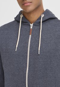 Un hoodie zippé gris foncé avec une texture douce, doté d'un cordon beige, d'une fermeture éclair en argent et d'une petite étiquette en cuir près du cou.