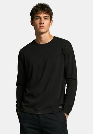 Jeune homme portant un t-shirt noir uni à manches longues et un pantalon foncé, debout avec les mains dans les poches devant un fond clair.