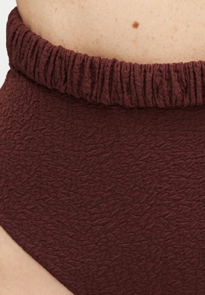 Gros plan sur un tissu texturé bordeaux avec une ceinture élastique portée sur le bas du torse, montrant un petit grain de beauté marron sur la peau au-dessus.