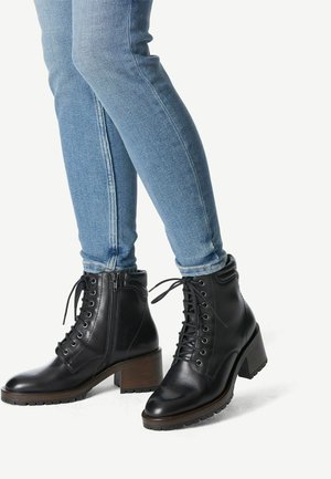 Botas de tobillo de cuero negro con un tacón grueso, cordones en la parte delantera y detalle de cremallera lateral, combinadas con jeans ajustados de color azul claro.