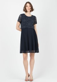 Marineblaues Kleid mit einem floralen Spitze-Oberteil und kurzen Ärmeln. Der Rock ist schwingend und halbtransparent, mit einer taillierten Silhouette unter der Spitze.