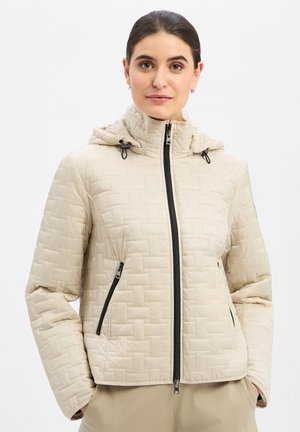 Frau mit dunklen Haaren, die eine beige gesteppte Jacke mit schwarzem Reißverschluss und Kapuze trägt, steht mit den Händen in den Taschen vor einem weißen Hintergrund.