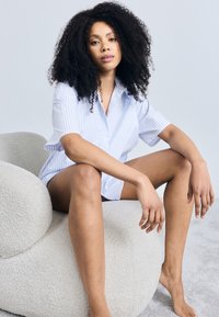 Πουκάμισο με κοντά μανίκια σε ανοιχτό γαλάζιο με ρίγες και άνετη εφαρμογή. Το μοντέλο κάθεται σε μια υφασμάτινη, στρογγυλή, εκρού καρέκλα με υφή.