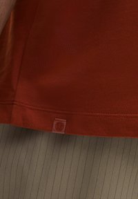 Camisa de algodón color óxido con un acabado texturizado y una pequeña etiqueta de logo en el dobladillo, superpuesta sobre una tela verde olivo a rayas.