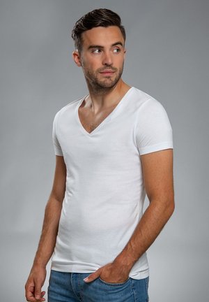 Witte V-hals t-shirt van zachte katoen, korte mouwen, aansluitend ontwerp, gedragen met blauwe denimjeans, geposeerd tegen een grijze achtergrond.