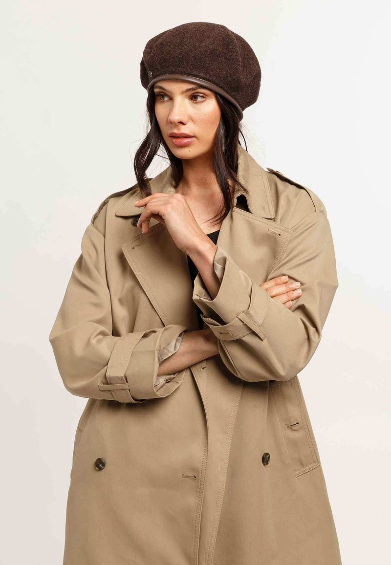 Bež trench coat lõdva lõikega, topeltkinnitatud nööbid ja laiad pöidlad; pruun villane barett nahkrihmaga.