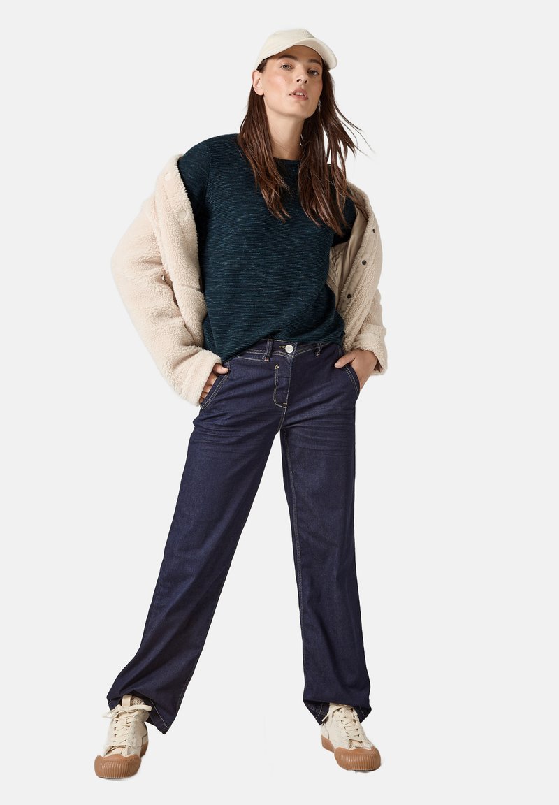 Dunkelblaue Denim-Jeans mit lockerer Passform, kombiniert mit einem strukturierten dunklen Pullover und einer beigen Fleecejacke, ergänzt durch beige Sneakers und eine Mütze.