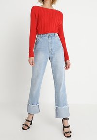 Haut rouge à manches longues côtelé, jeans bleu clair taille haute avec ourlet effiloché, et sandales à talons noires avec accents cloutés.