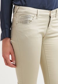 Beige jeans med detaljer av nitar på fickorna, tillverkade av denim med en slät yta. Dragkedja och knappstängning, figurnära design.