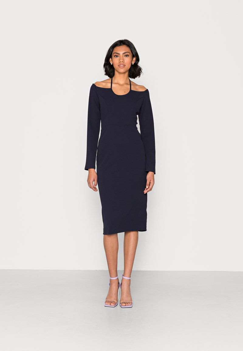 Vestido de manga longa azul-marinho com design off shoulder, silhueta ajustada, comprimento até a metade da panturrilha e textura suave; combinado com sapatos de salto com tira no tornozelo em lilás claro.