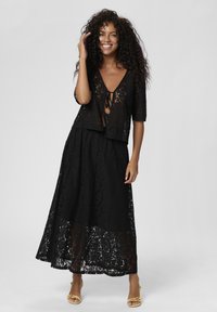 Schwarzes Spitzen-Crop-Top mit kurzen Ärmeln, tiefem V-Ausschnitt und Bindedetail; langer schwarzer Spitzenrock mit fließendem Design und transparentem unteren Teil.
