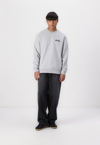 Gray sweatshirt with round neckline and ribbed cuffs, featuring a black logo on the chest, paired with loose-fitting dark jeans and black sneakers.