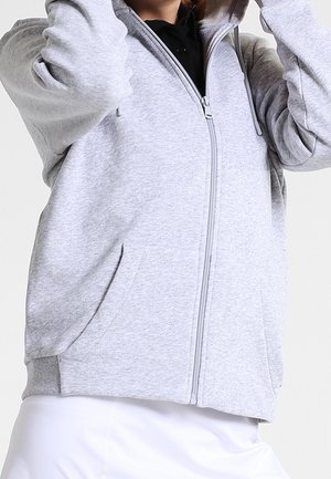 Personne portant un sweat à capuche gris clair zippé avec des poches sur un t-shirt noir, posant avec les bras levés et les mains près de la tête.