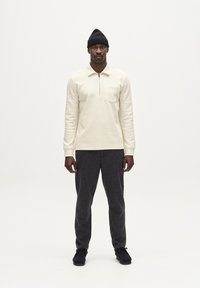 Krémszínű, hosszú ujjú póló negyed cipzárral és zsebbel a mellkasán, sötétszürke, texturált nadrággal és fekete cipővel párosítva. Sapka a fejen.