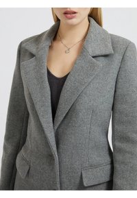 Blazer gris de lana con solapa muesca, dos bolsillos frontales y una textura en la tela. Presenta una camiseta gris superpuesta debajo.
