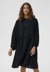 Rochie neagră cu mâneci lungi și un decolteu butonat. Prezintă broderie texturată pe piept și o siluetă lejeră. Material moale.