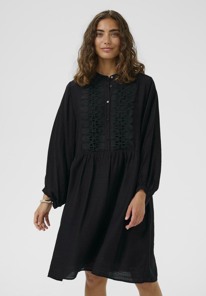 Rochie neagră cu mâneci lungi și un decolteu butonat. Prezintă broderie texturată pe piept și o siluetă lejeră. Material moale.