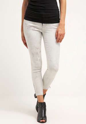 Femme portant un jean skinny gris clair avec un motif marbré subtil, un top noir sans manches et des bottines noires à bouts ouverts, debout jambes croisées.
