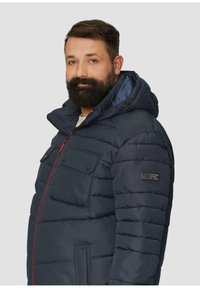 Marineblaue Steppjacke mit Kapuze, horizontalen Nähten, zwei Taschen auf der Brust mit Druckknöpfen und roten Reißverschlussakzenten. Weicher Stoff.