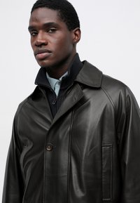 Manteau en cuir marron foncé avec un col classique, une fermeture à un bouton et une poche latérale, offrant une finition texturée et une apparence lisse.