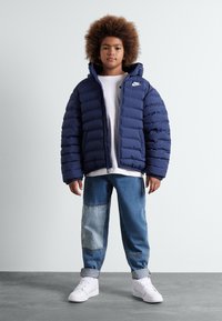 Giubbotto imbottito nero con cuciture orizzontali, dotato di cappuccio e chiusura con zip. Abbinato a pantaloni in denim azzurro chiaro e sneakers bianche.