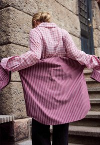 Langes Karohemd mit pink-weißer Karomuster auf der Vorderseite und vertikalen pink-weißen Streifen auf der Rückseite. Weicher Stoff, lockere Passform.