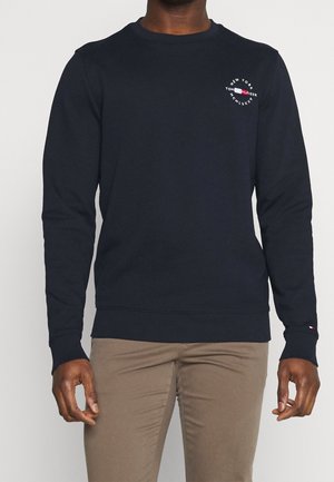 Mężczyzna ubrany w granatową długą bluzę z małym okrągłym logo Tommy Hilfiger na piersi i beżowe spodnie.
