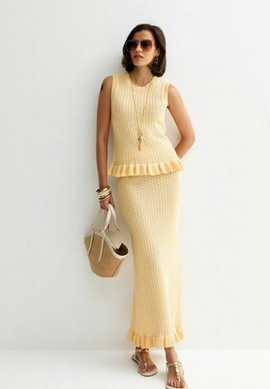 Mujer con un top sin mangas de punto amarillo y una falda larga a juego, gafas de sol, sandalias, pulseras, collar y sosteniendo un bolso beige.