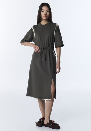 Kobieta w oliwkowo-zielonej sukience midi z bocznym rozcięciem i wiązanym paskiem w talii, w połączeniu z brązowymi wsuwanymi butami zdobionymi frędzlami.