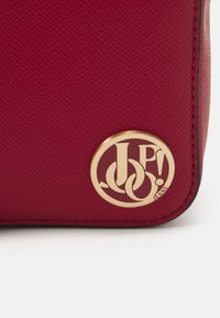 Un coin de sac en cuir synthétique rouge présentant un logo circulaire en métal doré embossé avec "JP! JEANS" et des lettres entrelacées. Détails de surface texturée.