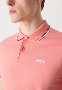 Chemise polo rose en coton avec patte de boutonnage à deux boutons, col à rayures contrastantes et détail brodé "Barbour International".