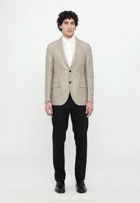 Hellbraun karierter Blazer mit einreihigem Design, zwei schwarzen Knöpfen, kombiniert mit einem weißen Hemd und schwarzen Hosen, abgerundet mit schwarzen Schuhen.