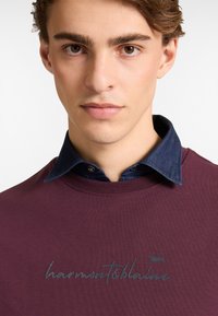 Bukserna dukserica boje burgundac s plavim denimski ovratnikom, s zelenim izvezenim natpisom "harmont&blaine" preko prsiju.