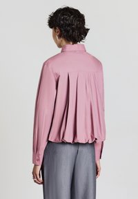 Persona che indossa una camicia a maniche lunghe rosa a vestibilità ampia con pieghe sulla schiena e pantaloni grigi a gamba larga, in piedi contro uno sfondo neutro.
