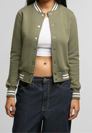 Personne portant une veste universitaire vert olive avec des bandes blanches aux poignets et au col, sur un crop top blanc et un jean bleu foncé.