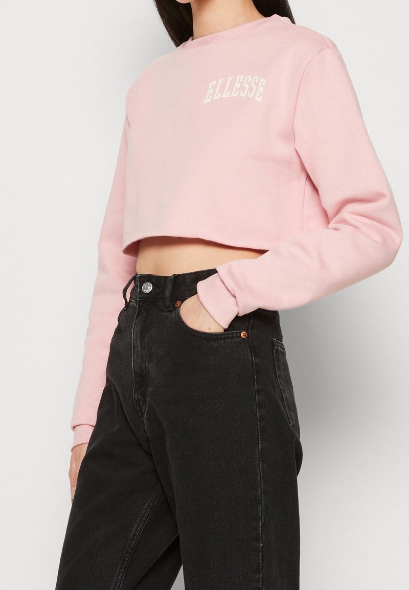 Felpa corta rosa con maniche lunghe, con il logo "ELLESSE" bianco. Abbinata a jeans neri in denim a vita alta.
