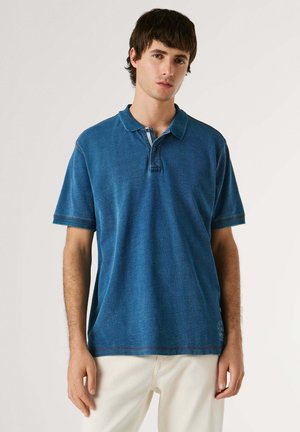 Joven con cabello corto y oscuro, vistiendo una camiseta tipo polo azul de manga corta y pantalones blancos, sobre un fondo claro y sencillo.