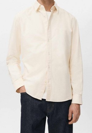 Homme portant une chemise crème à manches longues boutonnée par-dessus un sous-vêtement blanc avec un jean bleu foncé, une main dans la poche.