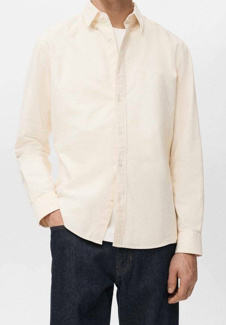 Homme portant une chemise crème à manches longues boutonnée par-dessus un sous-vêtement blanc avec un jean bleu foncé, une main dans la poche.