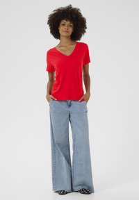 Piros v-nyakú póló, puha anyagból készült, széles szárú világoskék farmernadrággal párosítva. Az öltözet laza szabású és hétköznapi stílust képvisel.