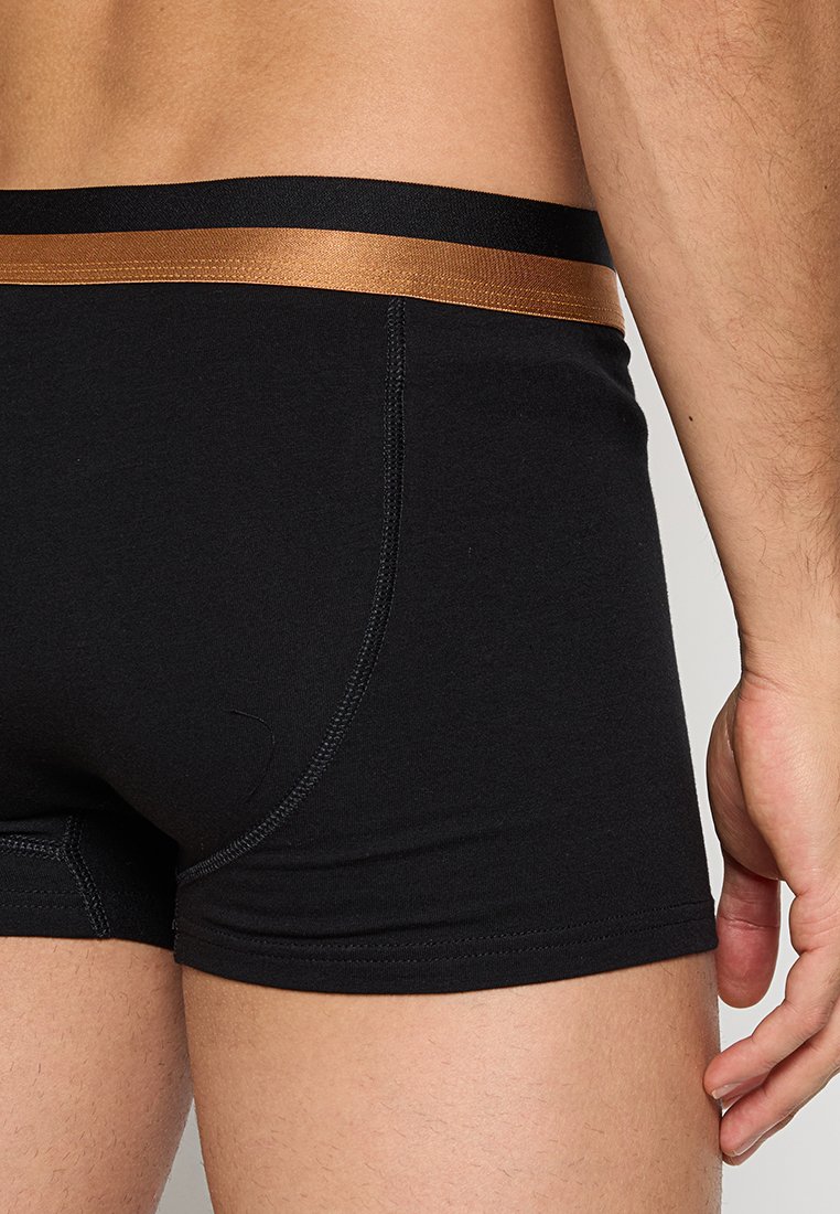 Schwarze Boxer-Briefs mit einer glatten Textur, ausgestattet mit einem kontrastierenden braunen elastischen Bund. Sichtbare Nähte an den Seiten.