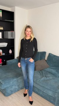 Γυναίκα με ξανθά μαλλιά που φοράει μαύρο πουκάμισο μακρυμάνικο, μπλε τζιν και μαύρα ψηλοτάκουνα, στέκεται σε σαλόνι κοντά σε μπλε καναπέ και βιβλιοθήκη.