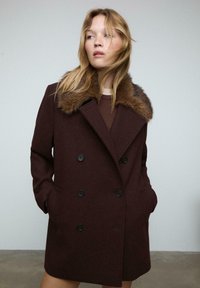 Giovane donna che indossa un cappotto doppiopetto marrone scuro con un ampio colletto di pelliccia, mani in tasca, che guarda di lato su uno sfondo neutro.