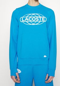 Felpa blu di Lacoste, con un grande logo bianco, collo a costine e tasca frontale. Tessuto morbido, maniche lunghe con polsini.