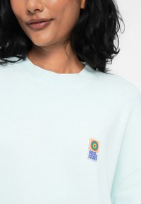 Šviesiai mėlynas megztinis su apvaliu iškirpimu ir minkšta tekstūra, ant krūtinės turi daugiakampį "Spalvų namų" logotipo pleistrą.