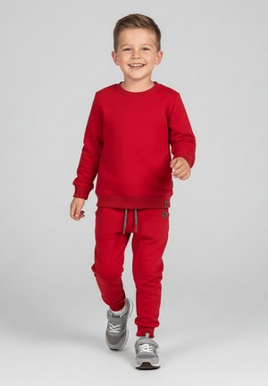 Uśmiechnięty chłopiec idący do przodu, ubrany w czerwoną bluzę z kapturem, pasujące czerwone spodnie dresowe i szare trampki na jednolitym jasnym tle.